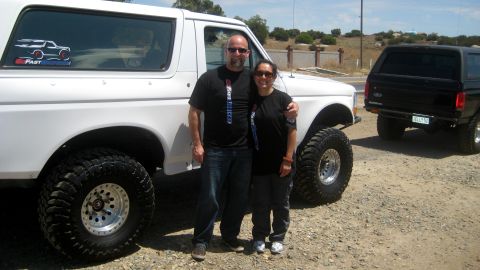 1996 Ford Bronco - Two Ford Broncos, a white one and a black one, parked outdoors with two people posing.