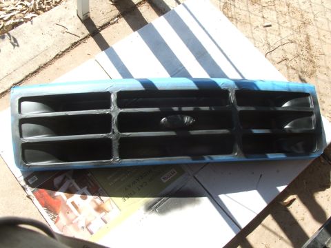 1995 Ford Bronco - A vehicle grille is prepped for painting, with blue tape outlining the edges and a central Ford emblem.