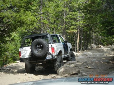 1995 Ford Bronco - White full-size Ford Bronco with the hardtop removed and a bikini/soft top over the front, flexing over a rocky shelf on a pine-lined mount…