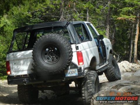 1995 Ford Bronco - White full-size Ford Bronco with the top removed and a black roll cage crawls over a rocky trail, viewed from the rear three-quarter with a…
