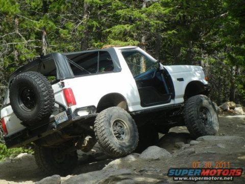 1995 Ford Bronco - A white full-size Ford Bronco with its hardtop removed and a black soft top over the cab crawls up granite ledges on a pine-forest trail, d…