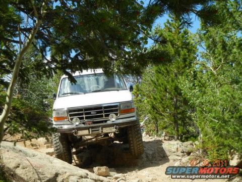 1995 Ford Bronco - A white Ford full-size 4x4 photographed head-on while crawling over rocks on a wooded trail, showing a lifted suspension, chrome front bump…