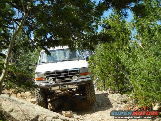 White lifted Ford 4x4 climbing rocks on a forested off-road trail, front view