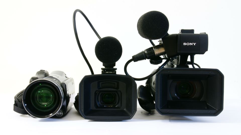 Three video cameras, two professional models with microphones and one smaller camcorder, arranged on a white surface