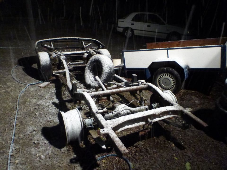 Car chassis covered in snow, with a trailer and another car in the background