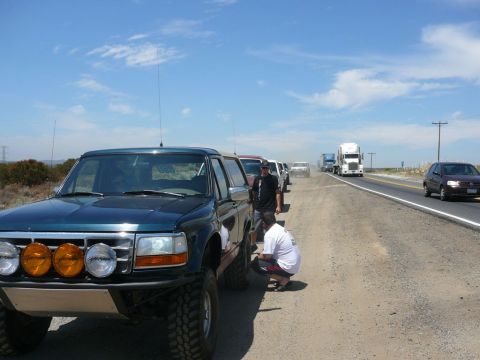 1988 Ford Bronco - A line of vehicles, including a modified Ford Bronco, are parked on the shoulder of a highway under a partly cloudy sky.