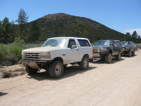 1988 Ford Bronco - A convoy of SUVs, led by a white 1992 Ford Bronco, travels on a dusty trail through a hilly, arid landscape.