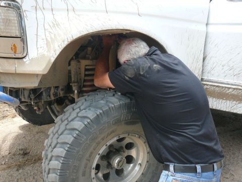 1988 Ford Bronco - Mechanic works on the front suspension of a white truck, with SUPERMOTORS.NET logo in the corner.