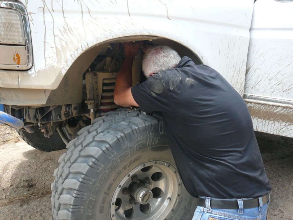 Man working on front suspension of a white truck with large off-road tires. SUPERMOTORS.NET logo visible