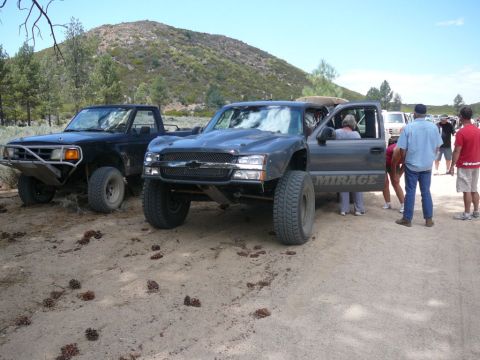 1988 Ford Bronco - Two modified trucks are parked on a dirt lot with a hilly, wooded background.