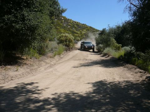 1988 Ford Bronco - A truck drives down a dusty dirt road surrounded by dry brush and trees under a clear blue sky.