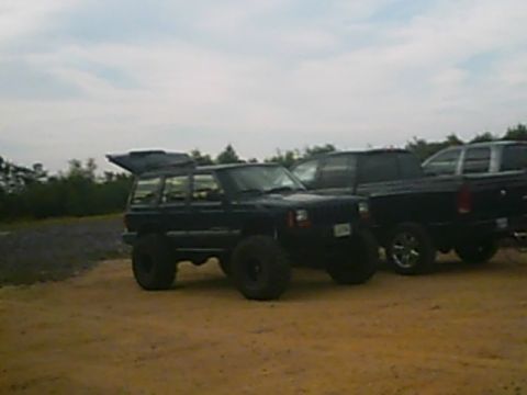 1988 Ford Bronco - A black Jeep Cherokee XJ is parked on a dirt lot next to a black pickup truck under a cloudy sky.
