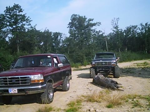 1988 Ford Bronco - Two Ford Broncos are parked on a dirt path surrounded by trees under a partly cloudy sky.