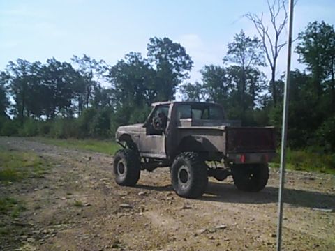 1988 Ford Bronco - A heavily modified pickup truck with oversized tires is seen driving on a dirt road through a wooded area.