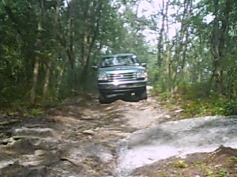 1988 Ford Bronco - A green SUV navigates a rough, unpaved path through a dense forest.