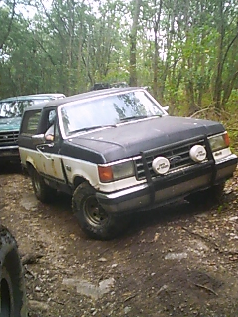 1988 Ford Bronco - A two-tone Ford Bronco navigates a muddy, wooded path with another vehicle visible in the background.