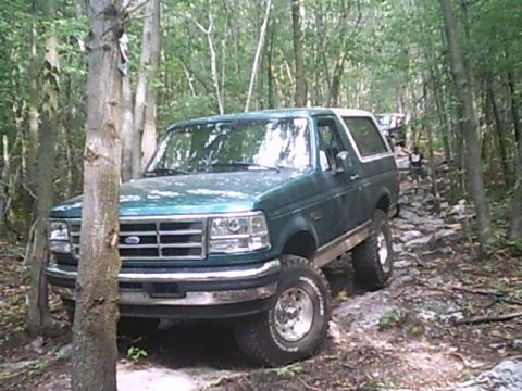 1988 Ford Bronco - A green Ford Bronco navigates a challenging rocky path through a wooded area, with another vehicle visible further down the trail.