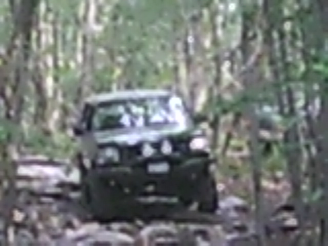 Dark SUV driving on a rocky, wooded trail. Lights are on the front