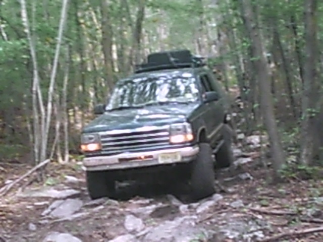 Green SUV with roof rack driving on a rocky, wooded trail. Grille suggests a 1992 model