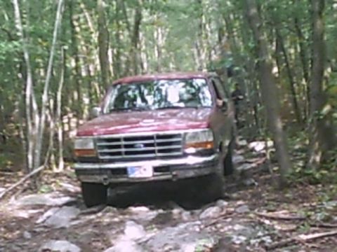 1988 Ford Bronco - A red SUV navigates a challenging, rocky path through a dense forest.