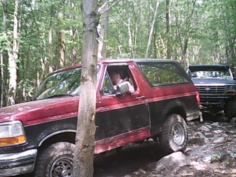 1988 Ford Bronco - Two Ford Broncos navigate a challenging, wooded off-road course with rocky terrain.