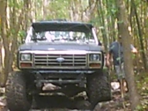 1988 Ford Bronco - A dark-colored Ford Bronco navigates a wooded trail, its large tires gripping the terrain.