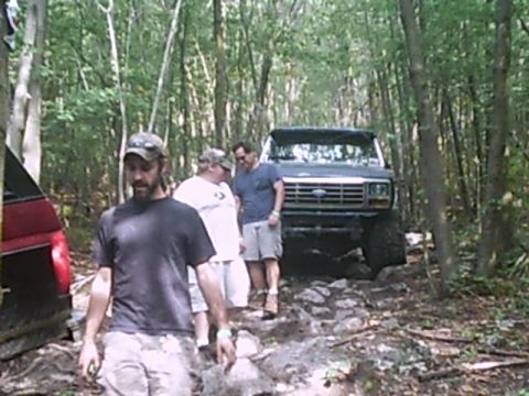 1988 Ford Bronco - A dark blue Ford truck navigates a rocky path through a wooded area with several people nearby.