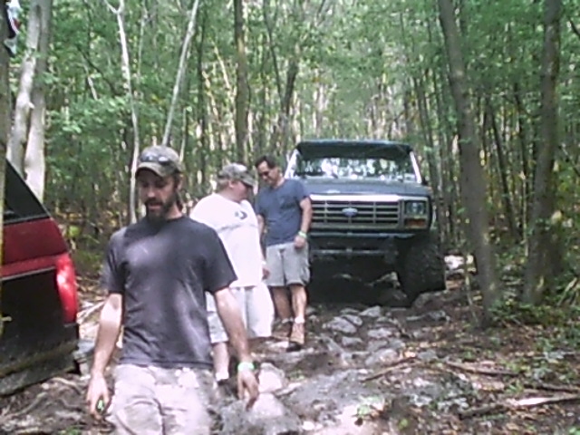 Dark blue Ford truck with a prominent grille and large tires on a rocky, wooded trail