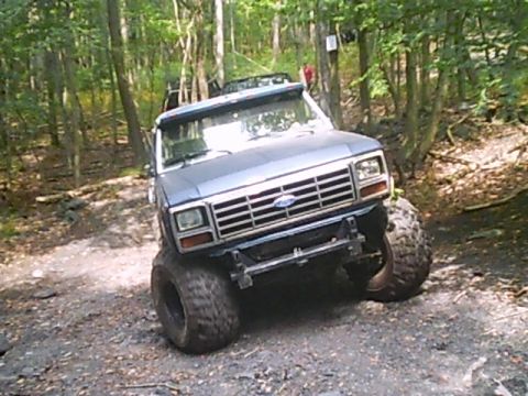 1988 Ford Bronco - Gray Ford Bronco with large tires navigates a wooded trail during an off-road event.