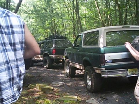 1988 Ford Bronco - A green Ford Bronco with a white top is parked on a dirt path, with another SUV behind it in a wooded area.