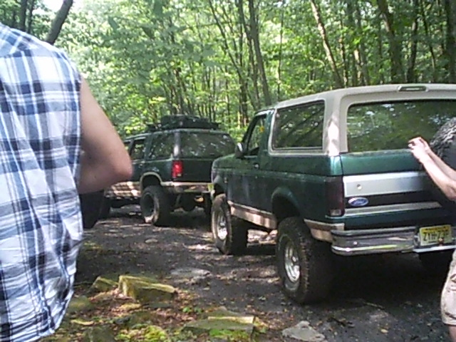 Green Ford Bronco with a white top, seen from the rear, parked on a dirt path in a wooded area