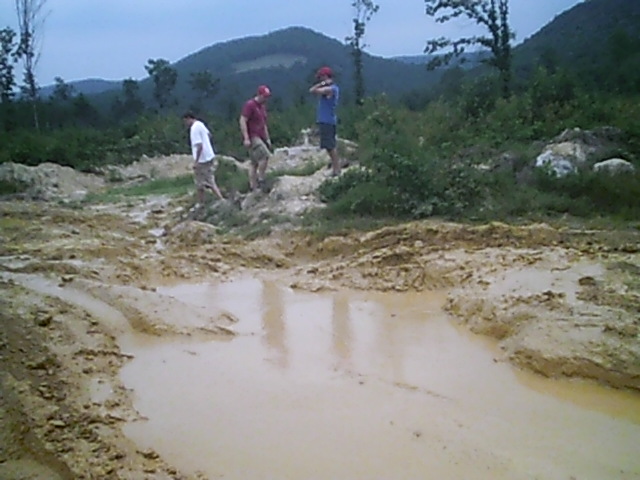 Three people walk through a muddy, off-road area with puddles and trees in the background. Watermark reads SUPERMOTORS.NET