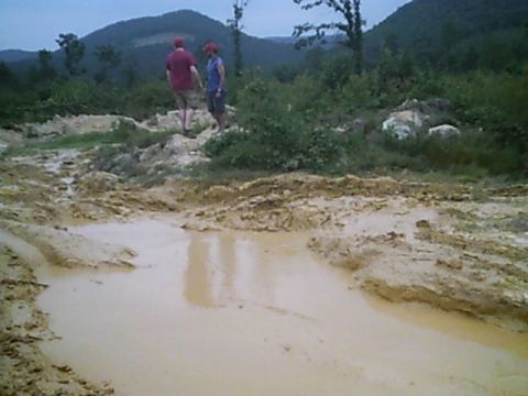 1988 Ford Bronco - Two men stand at the edge of a deep mud pit on a backcountry off-road trail with forested mountain ridges behind them.