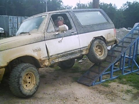 1988 Ford Bronco - This Ford Bronco is climbing a ramp, covered in mud. The driver is visible behind the wheel.