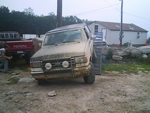 1988 Ford Bronco - A tan Ford Bronco is shown from the front, covered in mud, with a red Toyota truck in the background.