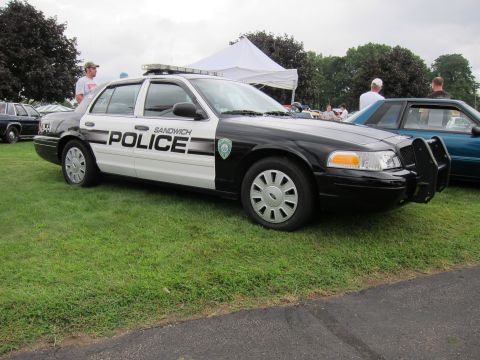 1991 Ford LTD Crown Victoria - Black-and-white Ford Crown Victoria marked SANDWICH POLICE with push bumper, parked on grass at a car show with onlookers.