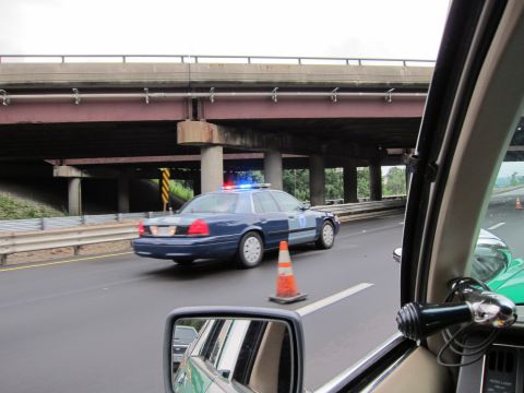 1991 Ford LTD Crown Victoria - blue police cruiser with lights drives along a highway beneath a concrete overpass, viewed from inside another vehicle.