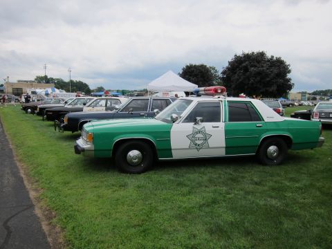 1991 Ford LTD Crown Victoria - Green-and-white Ford Sheriff sedan with star emblem at the head of a row of vintage cars on a grass show field.