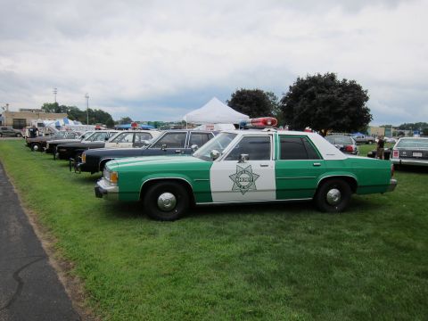 1991 Ford LTD Crown Victoria - Side view of a green-and-white Ford Sheriff sedan with light bar parked at the front of a line of show cars on grass.