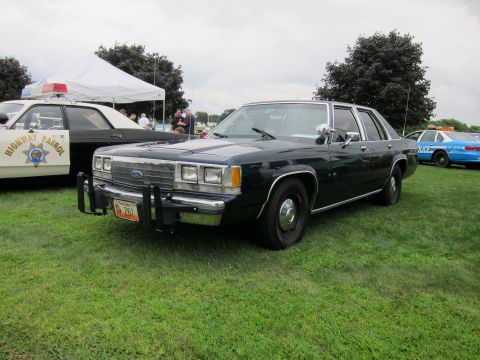1991 Ford LTD Crown Victoria - Dark-colored Ford sedan with chrome push bumper, front three-quarter view on grass with a Highway Patrol car alongside.