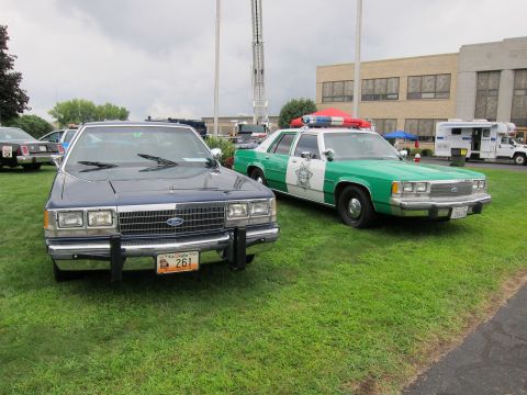 1991 Ford LTD Crown Victoria - blue Ford sedan with push bumper beside a green-and-white Sheriff cruiser on grass, building and trailers in background.