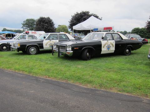 1991 Ford LTD Crown Victoria - Two black-and-white Highway Patrol sedans with door shields parked side by side on a grass show field near a paved edge.
