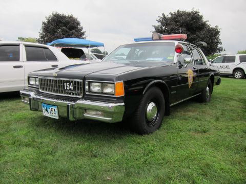 1991 Ford LTD Crown Victoria - Black police sedan number 154 with door shield and light bar, front three-quarter view on grass at an outdoor car show.