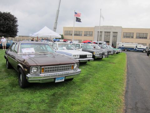 1991 Ford LTD Crown Victoria - long row of vintage police cars led by a brown sedan, parked on pavement before a building with US and POW-MIA flags.