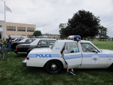 1991 Ford LTD Crown Victoria - white-and-blue marked police sedan numbered 839 sits on grass at an outdoor vehicle show, with a black car parked behind it.