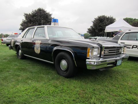 1991 Ford LTD Crown Victoria - black-and-white vintage police Ford sedan numbered 45 with a badge decal and single roof light sits on grass at a show.