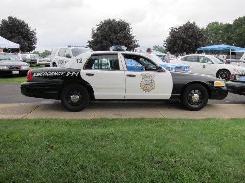 1991 Ford LTD Crown Victoria - Profile of a black-and-white police Ford Crown Victoria numbered 12 marked Emergency 9-1-1 with a badge decal on grass.