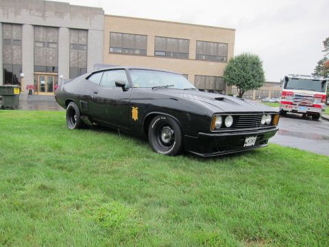 1991 Ford LTD Crown Victoria - matte black two-door muscle coupe with a gold star decal and rear spoiler sits on grass before a building and fire truck.