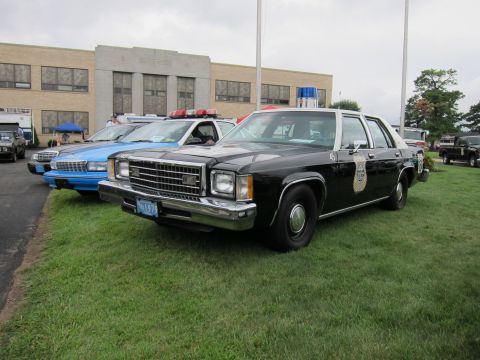 1991 Ford LTD Crown Victoria - black-and-white vintage police Ford LTD sedan with a badge decal and roof lights sits on grass, a blue sedan beside it.