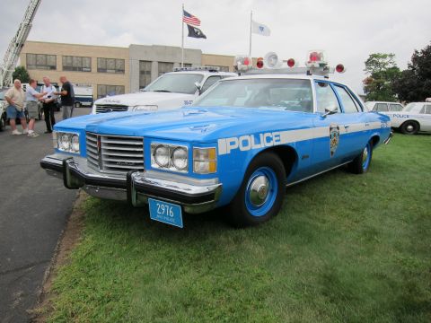 1991 Ford LTD Crown Victoria - blue 1970s NYPD Pontiac police sedan numbered 2916 with roof beacons sits on grass as spectators gather behind it.